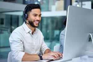 Happy young male sales agent talking to a customer via headset and working on computer station